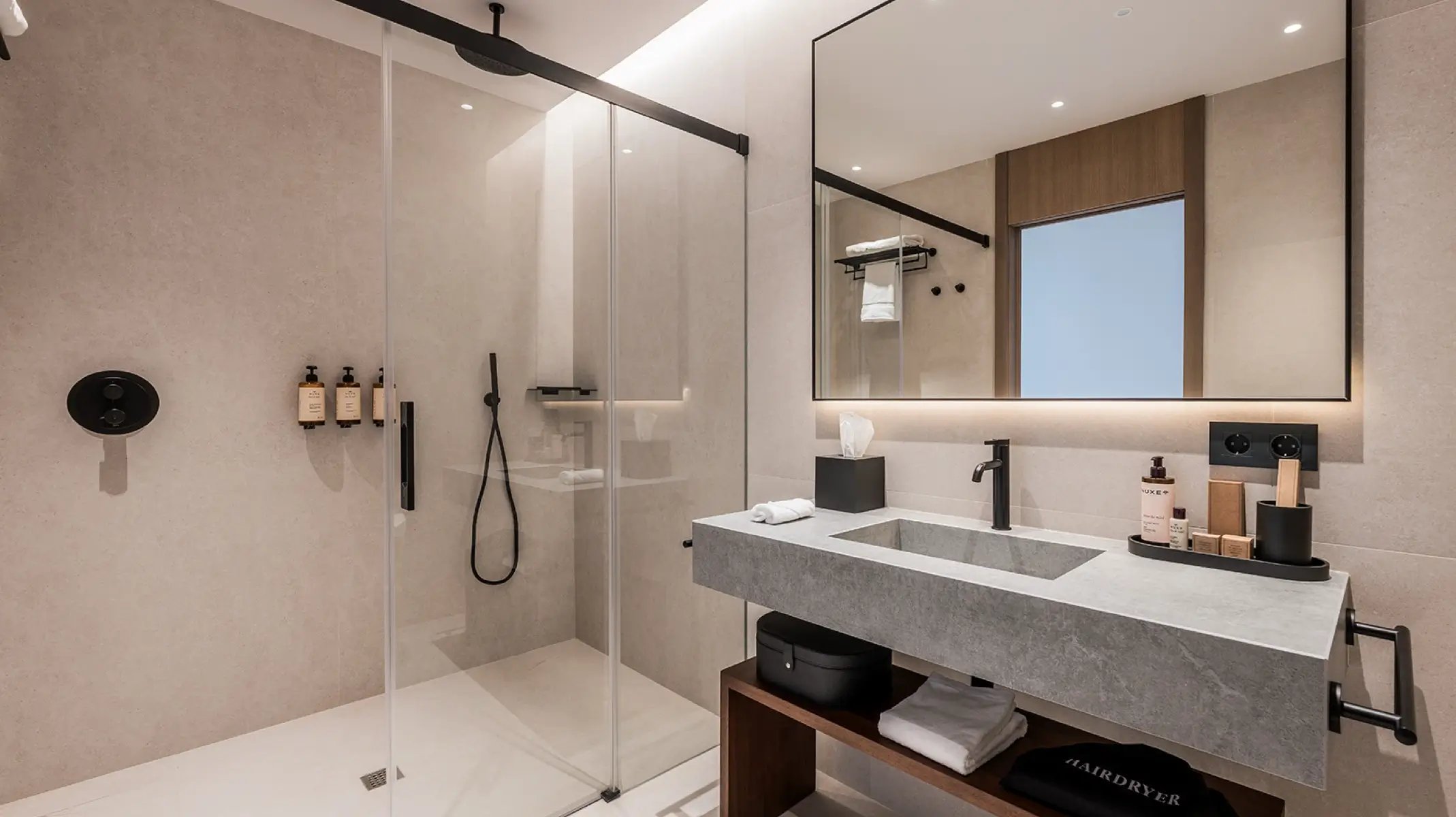 Baño de huéspedes de Naghoi — acabados en piedra de alta calidad, ducha de cristal y amenities de lujo Nuxe en una habitación de recuperación postquirúrgica en Marbella.